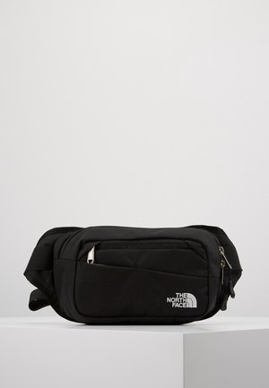 Sac banane noir The North Face avec plusieurs compartiments zippés et sangle réglable, présenté sur un socle blanc.