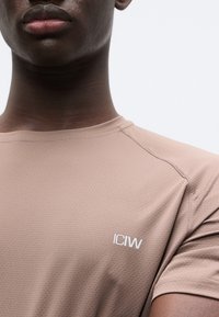 Ανοιχτό καφέ αθλητική μπλούζα με υφή, λαιμόκοψη και κοντά μανίκια. Διαθέτει λευκό λογότυπο "ICIW" στο αριστερό στήθος.