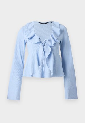 Blouse à manches longues bleu clair avec tissu texturé, col en V volanté et détail noué à l'avant, présentée sur un fond uni.