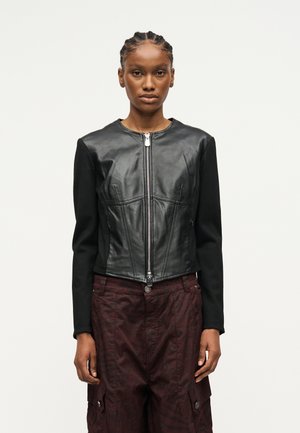 Γυναίκα με πλεξούδες, φοράει μαύρο δερμάτινο τζάκετ με φερμουάρ και σκούρο μπορντό παντελόνι cargo, στέκεται μπροστά σε απλό φόντο.