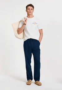 Balti medvilninė marškinėliai su mėlynu grafiniu piešiniu, derinami su tamsiai mėlynomis dryžuotomis kelnėmis. Modelis laiko kreminės spalvos rankinę ir dėvi brunzinio atspalvio sandalus.