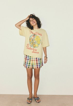 Sieviete ar cirtainiem matiem stāv pret vienkrāsainu sienu, valkājot dzeltens t-kreklu ar grafisku attēlu ar austerēm un citronu, rūtainas šortus un saskaņotus rūtainus sandales.