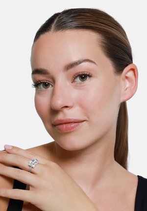 Kobieta z gładką skórą i włosami zaczesanymi do tyłu pokazuje srebrny pierścionek z trzema dużymi prostokątnymi, przejrzystymi kamieniami na lewej ręce.