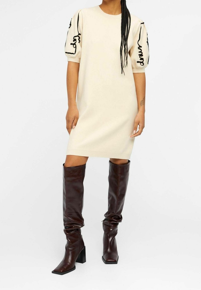 Vestido de punto color crema con mangas abullonadas cortas que presenta bordados abstractos en negro. Combinado con botas altas de cuero marrón oscuro.
