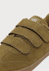 Chaussure en daim marron avec deux larges bandes velcro et des bords festonnés, avec une couture fine et de petites perforations sur le côté.
