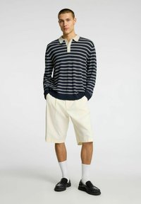 Polo a righe navy e bianche con maniche lunghe e colletto crema, abbinato a pantaloni corti crema fino al ginocchio e mocassini neri, con calze bianche in vista.