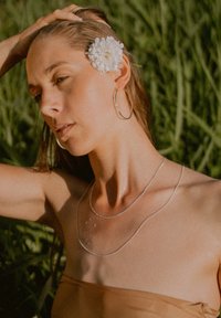 Silver necklaces layered on a bare shoulder, with water droplets visible. A large white flower accentuates the hairstyle against green foliage.