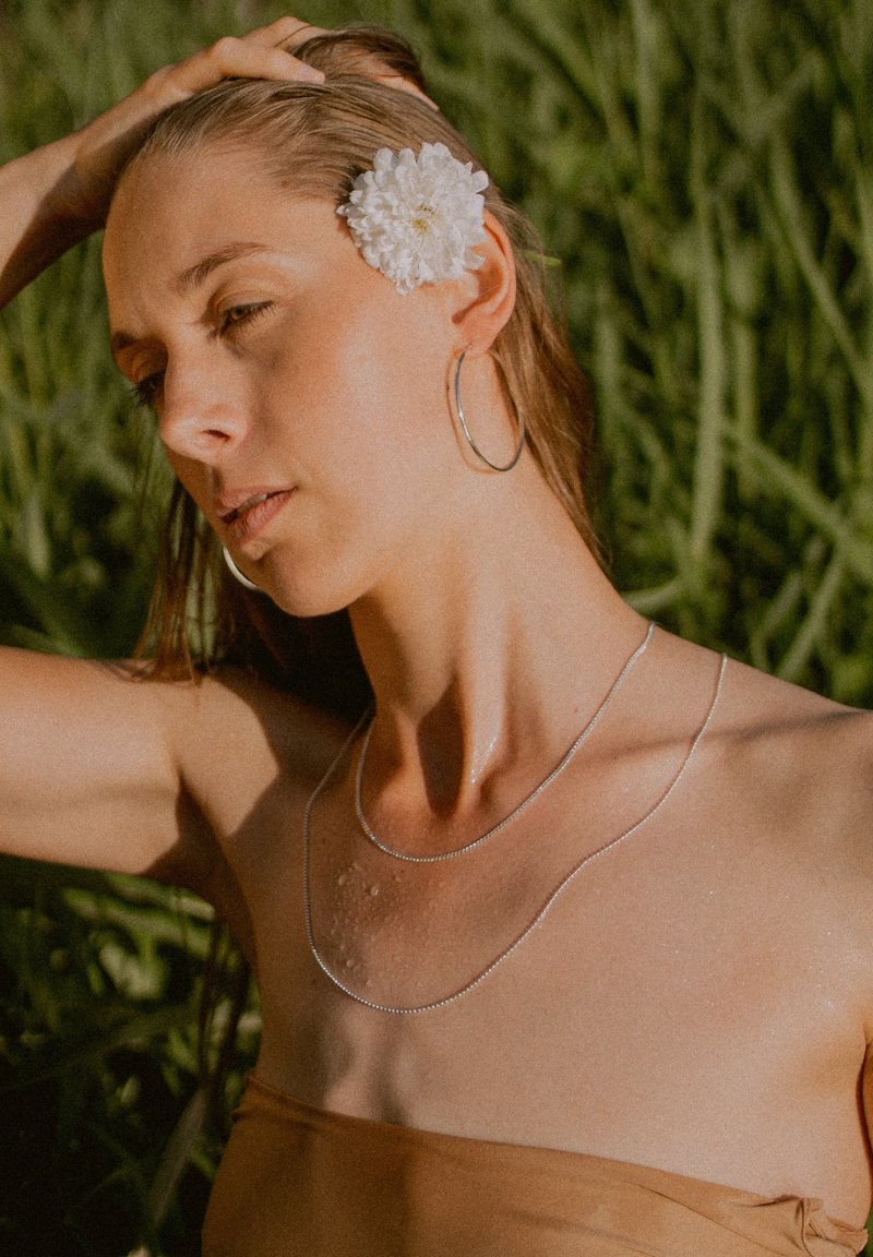 Silver necklaces layered on a bare shoulder, with water droplets visible. A large white flower accentuates the hairstyle against green foliage.