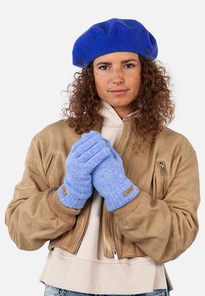 Blue knitted gloves with ribbed texture and a tan leather accent. Worn by a person in a beige bomber jacket and blue beret.