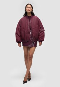 Vinröd bomberjacka med oversized passform, ribbade muddar och dragkedja; kombinerad med en glansig minikjol i matchande färg och spetsiga klackar.