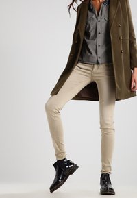 Olivově zelený kabát s dvouřadým zapínáním, šedá košile s vzorovaným límcem, béžové skinny džíny a černé lakované kožené kotníkové boty.
