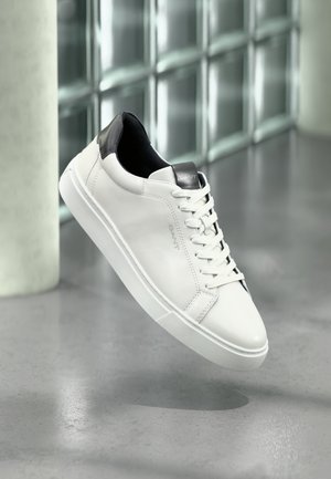 Tenis Alb din piele cu accente negre pe călcâi, bot rotund și talpă de cauciuc texturată. Prezintă șireturi plate și branding pe limbă.