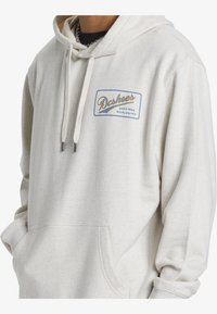 Cream-colored hoodie with a front pocket, drawstrings, and a logo patch in blue and gold. Made of soft fabric with a relaxed fit.