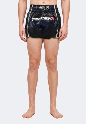 Homme portant un short de sport noir de la marque Venum avec le logo "TEKKEN 8" à l'avant, debout pieds nus devant un fond clair uni.