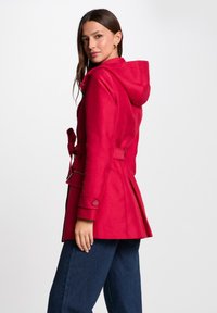 Cappotto rosso con cappuccio e vita fasciata, dotato di tasche con zip e fondo svasato. Texture liscia con un design pulito e senza motivi visibili.