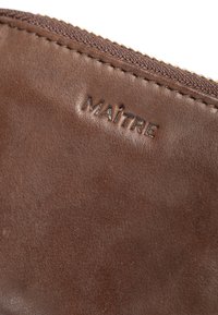 Portafoglio in pelle marrone con una superficie liscia, caratterizzato da un sottile logo in rilievo "MAÎTRE". Zip dorata con bordo in tessuto scuro.
