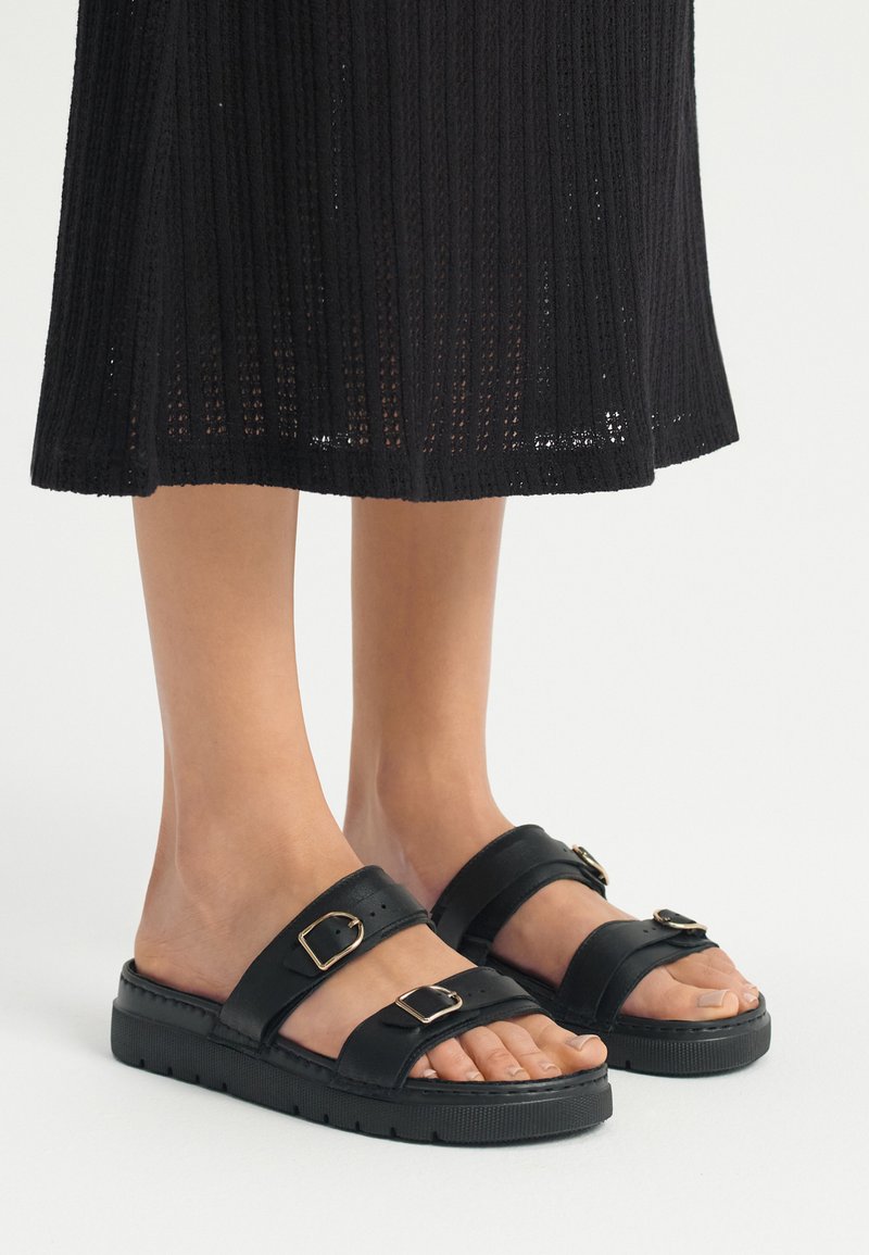 Czarne skórzane sandały z dwoma szerokimi paskami z klamrami, teksturowaną gumową podeszwą i otwartym noskiem, pokazane z czarną spódnicą o prążkowanej strukturze.