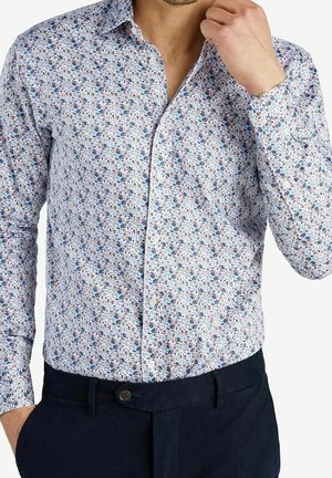 Homme portant une chemise blanche à manches longues avec un petit motif floral et un pantalon bleu foncé, main touchant le col.
