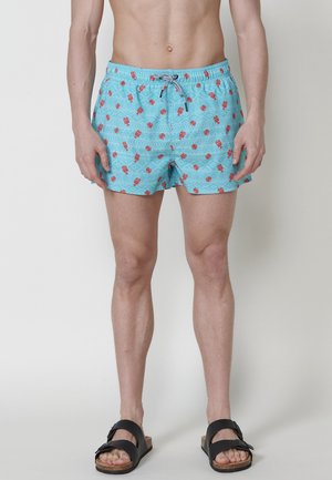 Pantaloni da bagno azzurri chiari con stampa di aragosta e cordoncino nero, realizzati in tessuto testurizzato, di lunghezza sopra il ginocchio, abbinati a sandali neri.
