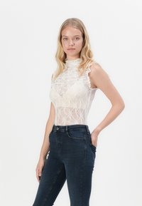 Lace bodysuit i krem med høy hals og ermeløs design, med bølgete kanter og en tett silhuett, kombinert med mørke skinny jeans.