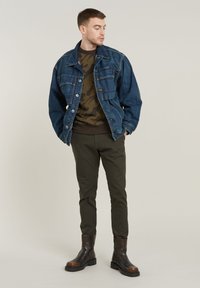 Chaqueta de denim con dos bolsillos en el pecho, sudadera de cuello redondo de camuflaje marrón, pantalones tapered en verde olivo y botas Chelsea de ante marrón oscuro.
