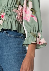 Blusa floreale verde con vita arricciata e maniche a volant, decorata con motivi di fiori rosa. Abbinata a jeans in denim blu.