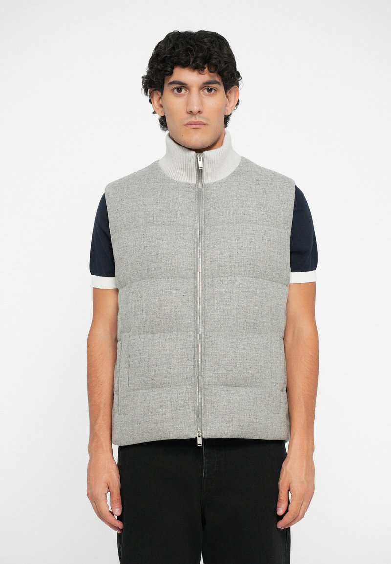 Vest puffer gri cu guler înalt, închidere cu fermoar și buzunare laterale; dispune de un material texturat cu un aspect matlasat. Puratat peste o cămașă bleumarin.