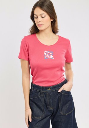 Femme portant une chemise rose à manches courtes avec un motif floral abstrait bleu et blanc, et un pantalon en jean foncé taille haute.