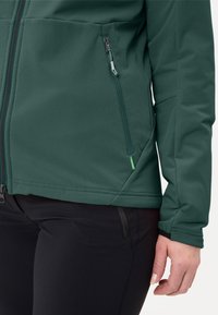 Giacca verde in soft-shell con tasca laterale con zip, superficie strutturata e design aderente. Pantaloni neri con un piccolo dettaglio con zip.