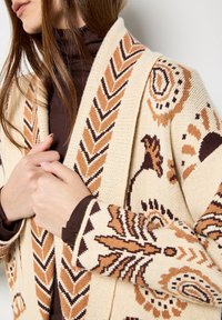 Beżowy sweter dzianinowy z tribalnymi wzorami w odcieniach brązu i pomarańczu, z narzutowym kołnierzem i otwartym przodem, podkreślający teksturę i szczegóły wzornictwa.