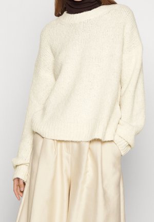 Pull en maille crème à coupe ample, avec des bords-côtes aux poignets et à l'ourlet. Porté par-dessus un col roulé noir. Associé à un pantalon en satin beige plissé.