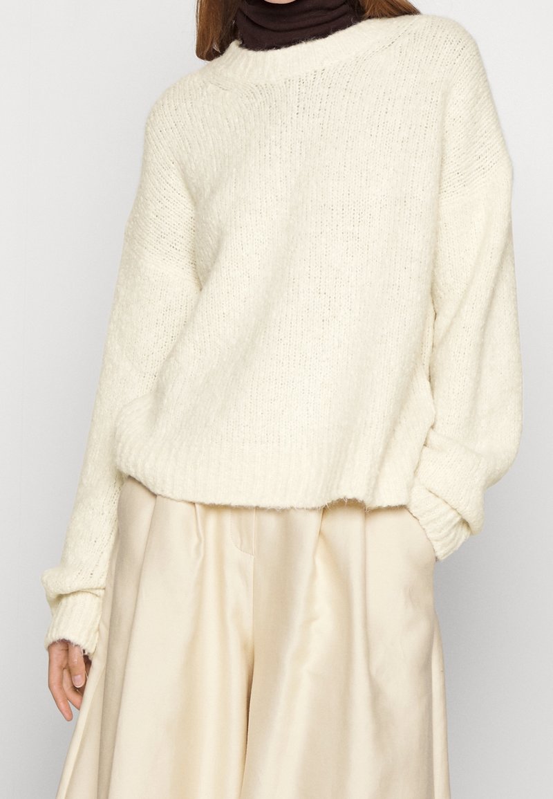Pull en maille crème à coupe ample, avec des bords-côtes aux poignets et à l'ourlet. Porté par-dessus un col roulé noir. Associé à un pantalon en satin beige plissé.