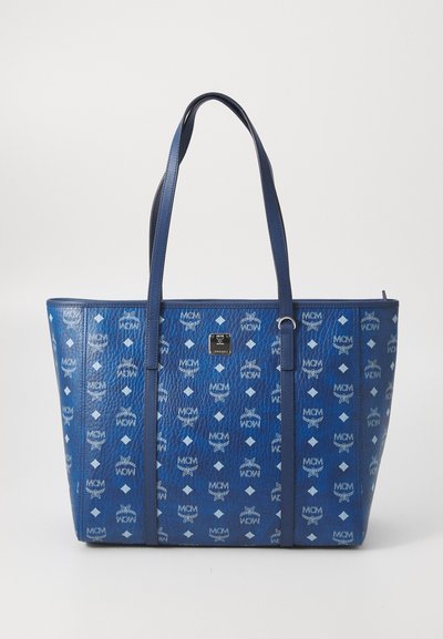 Blue leather tote bag with MCM logo pattern, double shoulder straps, and a small square metal brand plaque on the front center.