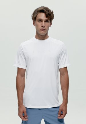 Muž so svetlo hnedými vlasmi, oblečený v jednoduchom bielom tričku s krátkym rukávom a svetlomodrých šortkách, stojaci pred svetlosivým pozadím.