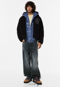 Czarna polarowa kurtka na niebieskiej bluzie z kapturem, jasna koszula w paski, szare szerokie dżinsy i brązowe buty z niebieskimi sznurówkami.