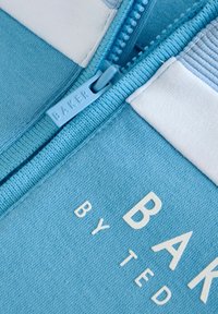 Svetlo modrá bundová mikina na zips s textúrovanou tkaninou, kontrastným bielym lemom a ťahákom na zips označeným "BAKER." Text je zobrazený v bielej farbe.