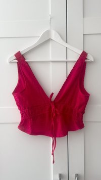 Rood, mouwloos crop top met een V-hals, gathered taille en een strikdetails. De stof lijkt getextureerd met een gegolfd patroon.