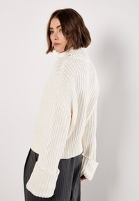 Maglione bianco a collo alto, con texture a coste, design cropped e maniche ampie. Indossato sopra pantaloni grigi a vita alta.