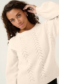 Ljusbeige stickad tröja med ribbad halsringning, med ett fiskbensmönster och ballongärmar för extra textur.
