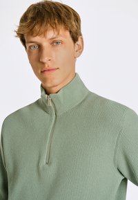 Ljust grön ribbad stickad tröja med hög krage och kort dragkedja. Har ett strukturerat tyg och en figurnära passform.