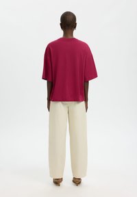 Άτομο που φοράει φαρδιά κόκκινη κοντομάνικη μπλούζα, φαρδιά μπεζ παντελόνια και καφέ σανδάλια με τακούνι, στέκεται με την πλάτη προς τα εμπρός σε απλό λευκό φόντο.