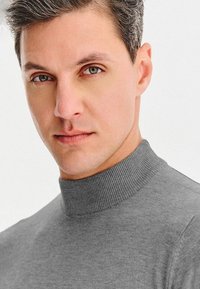 Szary, ryfrowany sweter z golfem o dopasowanym kroju, miękkiej fakturze i wysokim kołnierzu, noszony przez mężczyznę z krótkimi, jasnoszarymi włosami.