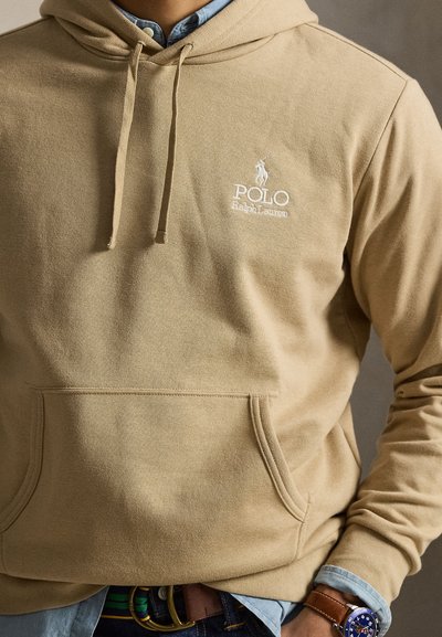 Hanorac bej Polo Ralph Lauren purtat peste o cămașă albastră, cu curea de piele maro și ceas albastru la încheietura mâinii stângi.