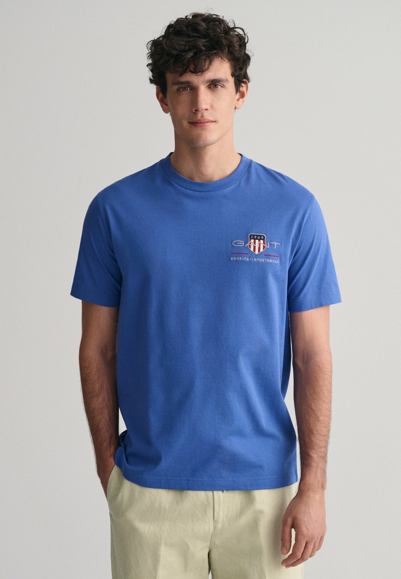 Modrá bavlněná tričko s krátkým rukávem, kulatým výstřihem a logem na hrudi. Tkanina vypadá hladce a má ležérní design.