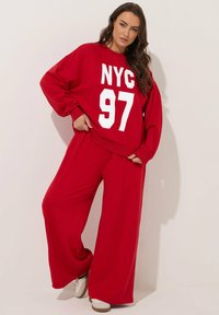 Sudadera roja con el texto blanco "NYC 97", combinada con pantalones anchos rojos a juego. Ambos hechos de un tejido suave y liso. Zapatillas blancas completan el look.