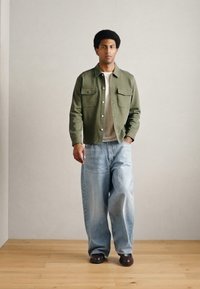 Giacca verde oliva con bottoni e due tasche sul petto sopra un maglione chiaro, abbinata a jeans di jeans azzurri a taglio ampio e scarpe scure.