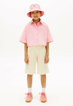 Dziecko stoi, ubrane w różowy kapelusz z szerokim rondem, różowo-białą koszulkę w paski z krótkim rękawem, kremowe bermudy, białe skarpetki i różowe trampki.
