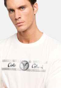 Witte crewneck t-shirt met een metallic zilveren grafische band met een circulair ontwerp en een Grieks sleutelpatroon. Zachte textuur, casual pasvorm.