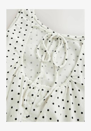 Blouse blanche sans manches à pois noirs, avec un col froncé et un nœud noué à l'avant.