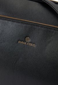 Schwarze Texturtasche mit goldfarbenem Reißverschluss und dem in Gold geprägten "ANNA FIELD"-Logo auf der Vorderseite. Glattes Stoffdesign mit dezenten Linien.